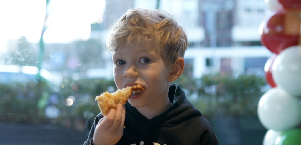 Pizza Enjoyment Image: Denis' Son Delights in a Slice at New York Pizza's Grand Opening.