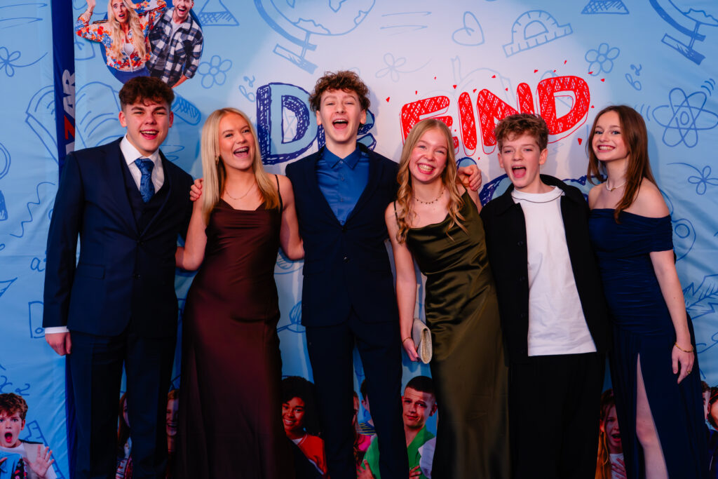 De jonge talenten van de cast van De Eindmusical poseren enthousiast voor de perswand tijdens de première van de theatershow, gefotografeerd door Monutes.