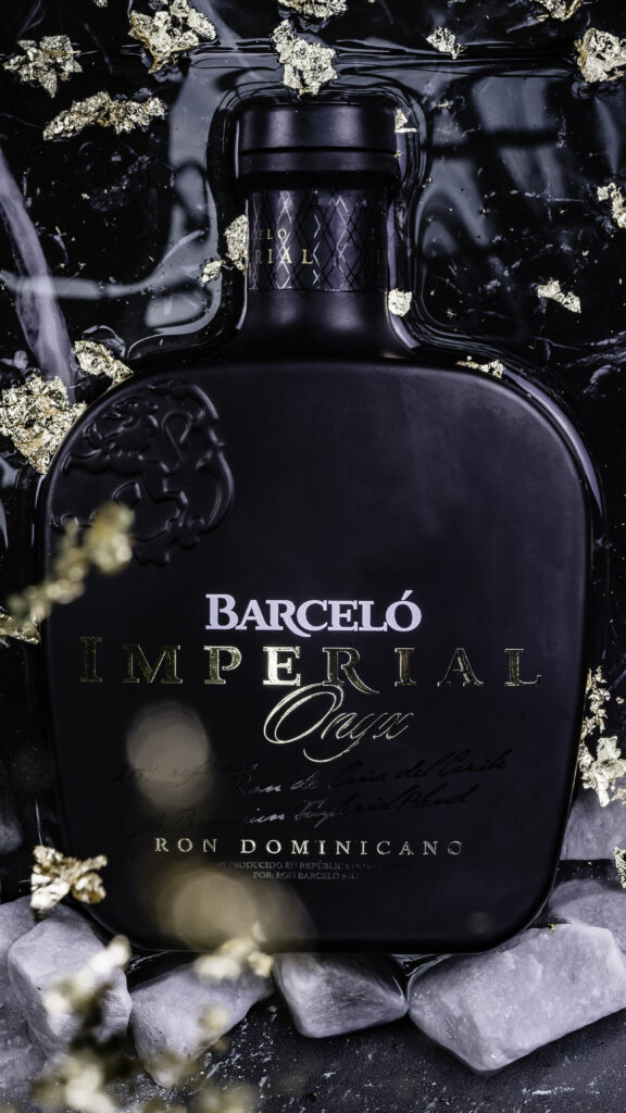 Luxe productfoto van een zwarte Barceló Imperial Onyx rumfles omringd door bladgoud en witte stenen in een professionele studioketting van Monutes.