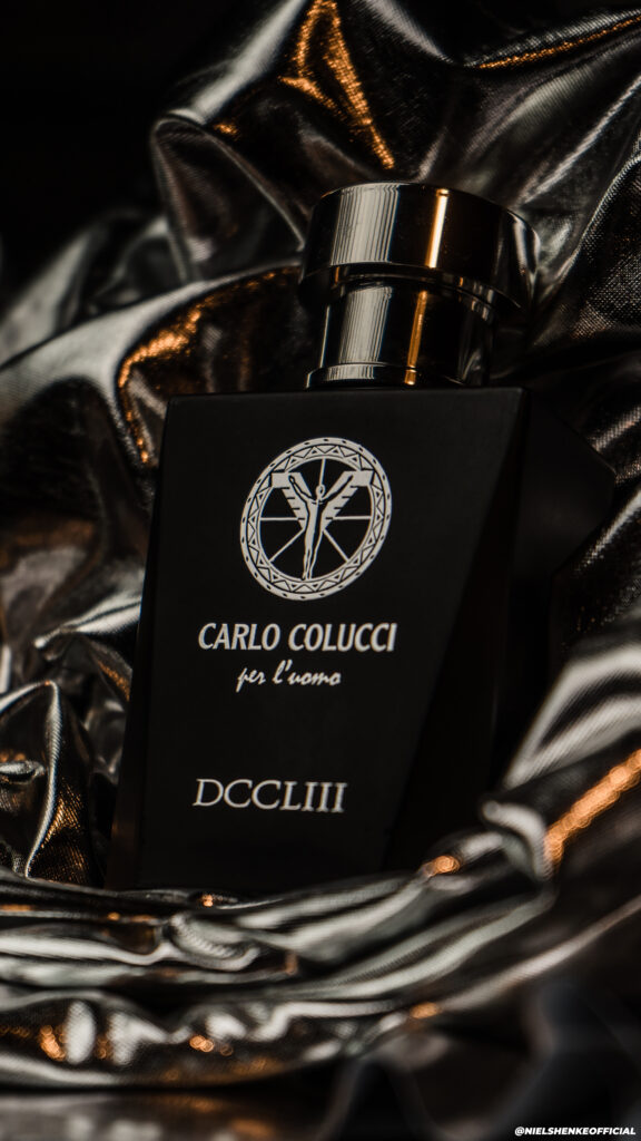 Een luxe productfoto van een matzwarte Carlo Colucci DCCLIII parfumfles tegen een achtergrond van glanzende, gedrapeerde zilveren stof, gefotografeerd door Monutes.