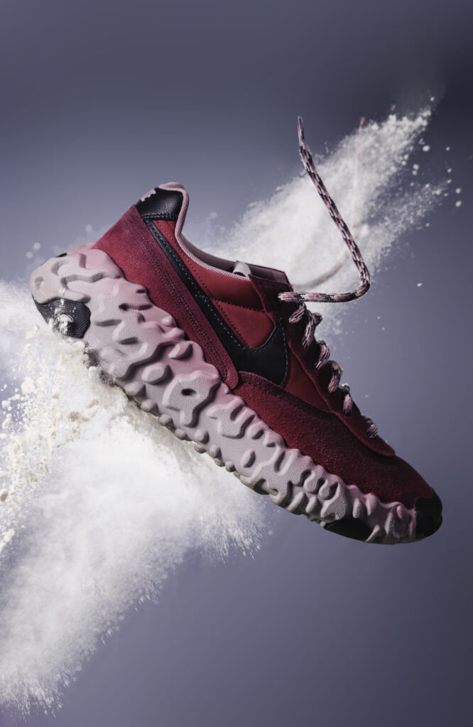 Een creatieve productfoto van een roze en magenta Nike Overbreak sneaker die door een witte poederexplosie vliegt, met zwevende veters tegen een strakke grijs-blauwe achtergrond, gefotografeerd door Monutes.