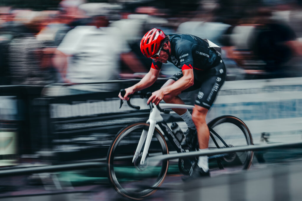 Een dynamische sportreportagefoto van een wielrenner met een rode helm in volle vaart, met een bewogen achtergrond die snelheid suggereert, gefotografeerd door Monutes.