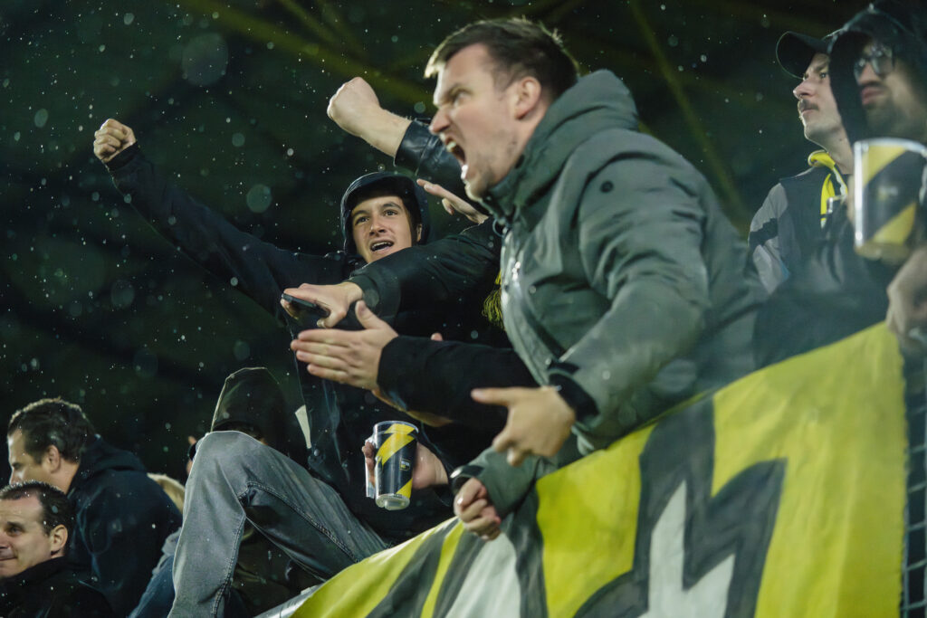 Een sfeervolle reportagefoto van juichende NAC Breda supporters in de regen tijdens een avondwedstrijd in het Rat Verlegh Stadion, gefotografeerd door Monutes.