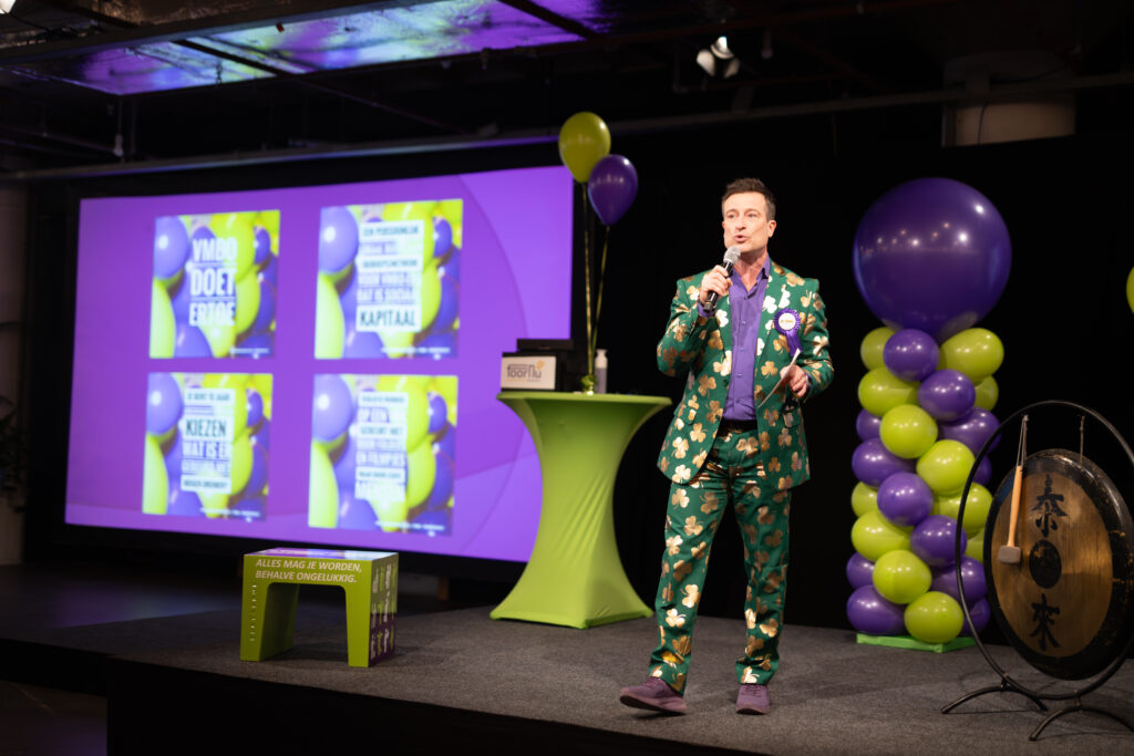 Een kleurrijke reportagefoto van MC Daniël Staakman in een groen pak met gouden klaverbladen op het podium van Amsterdam Nieuw-West On Stage, gefotografeerd door Monutes.