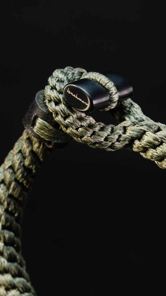 Een gedetailleerde macro-productfoto van een groen gevlochten Brahman armband met een zwart metalen sluiting voorzien van goudkleurige gravure, tegen een zwarte achtergrond, gefotografeerd door Monutes.