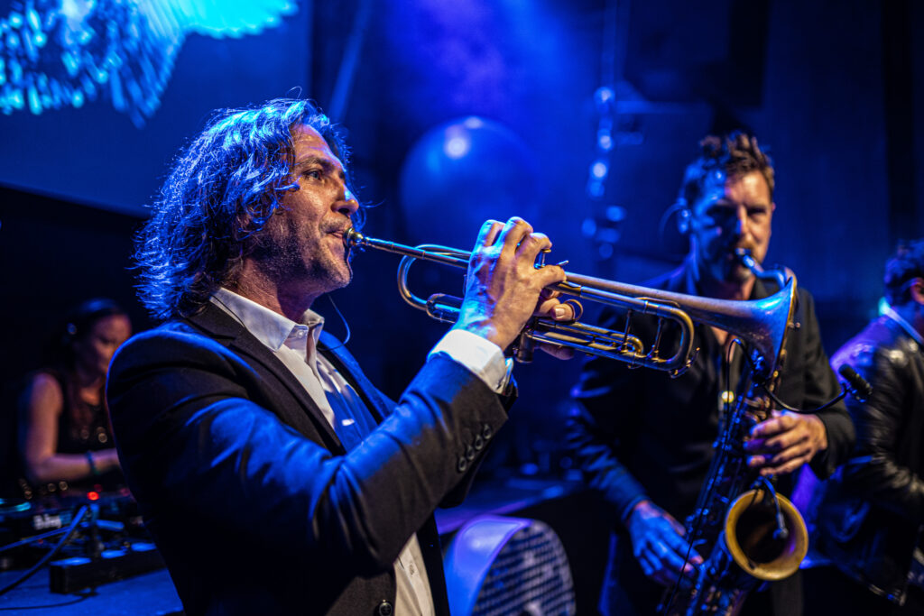 Een sfeervolle reportagefoto van een muzikant die trompet speelt tijdens een live optreden op een evenement, met een saxofonist op de achtergrond en blauwe podiumverlichting, gefotografeerd door Monutes.