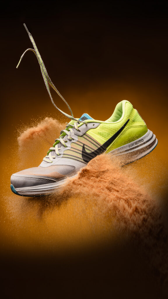 Een dynamische productfoto van een zwevende Nike hardloopschoen (neon geel, grijs en wit) met veters die omhoog zweven en opspattend zand op een warme oranje-bruine achtergrond, gefotografeerd door Monutes.