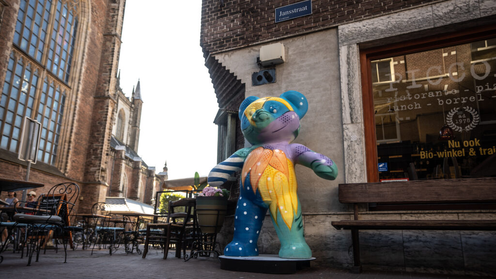 Een kleurrijke, beschilderde beer van het Robijn Robanksy project in de Jansstraat in Haarlem, met de Grote Kerk op de achtergrond, gefotografeerd door Monutes.