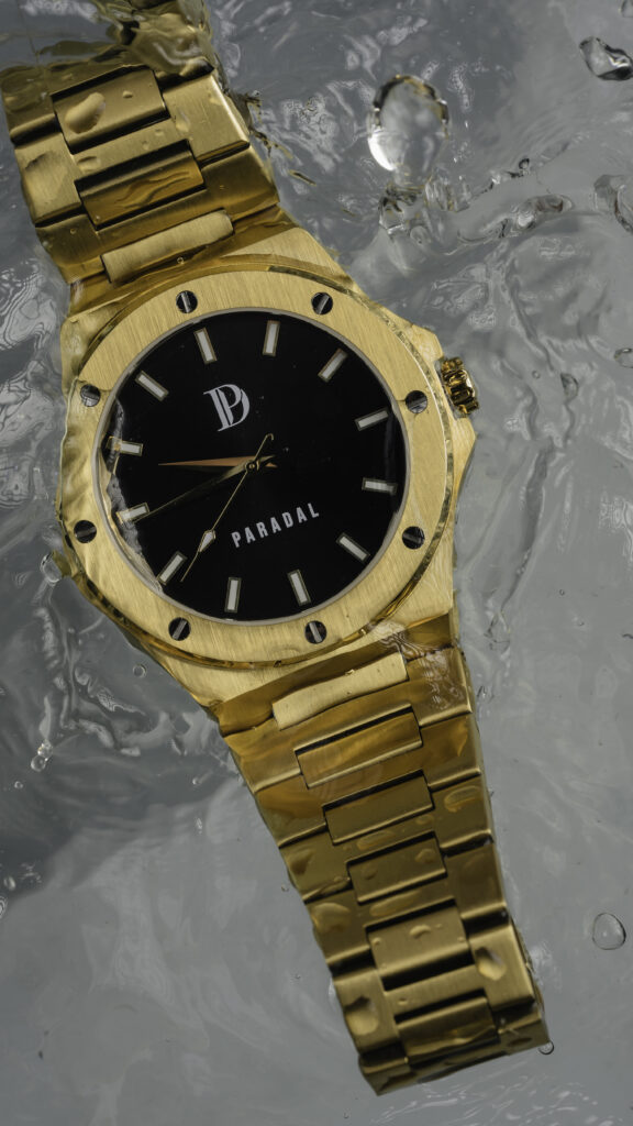 Een close-up productfoto van een gouden Paradal horloge met een zwarte wijzerplaat, liggend in bewegend water met opspattende druppels, gefotografeerd door Monutes.